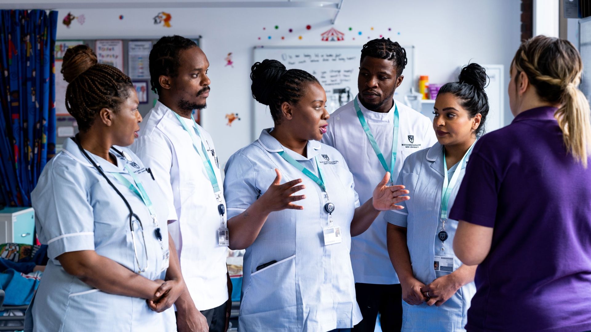 Five student nurses in uniform stood in discussion whilst facing a lecturer in a simulation ward